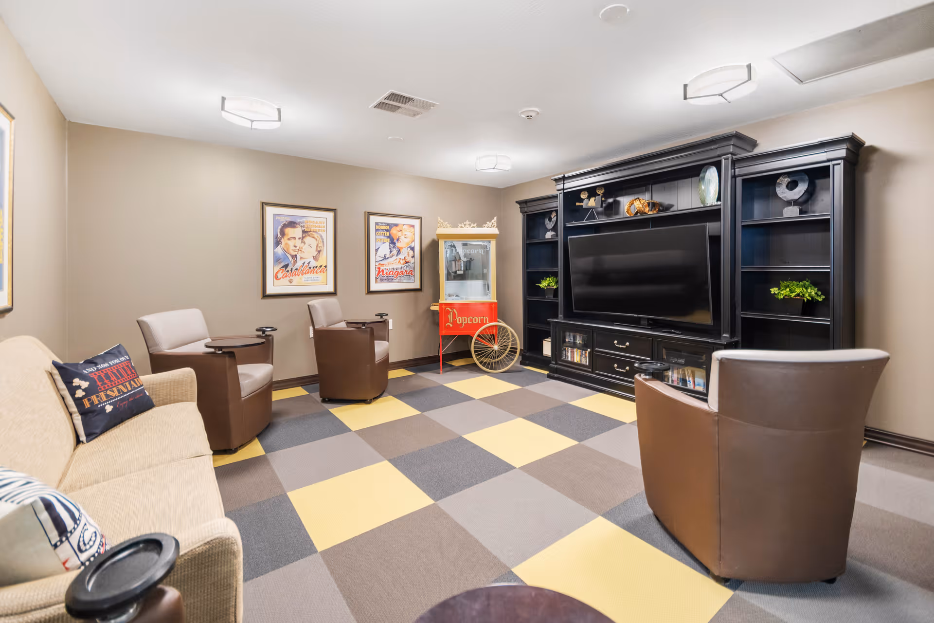 A cozy entertainment room with a large flat-screen TV set in a black wooden entertainment center. The room has a checkered carpet with yellow, gray, and brown squares. There are three brown leather chairs and a beige sofa with decorative pillows. On the wall, there are framed vintage movie posters and a red popcorn machine. The walls are painted beige and the ceiling has modern light fixtures.