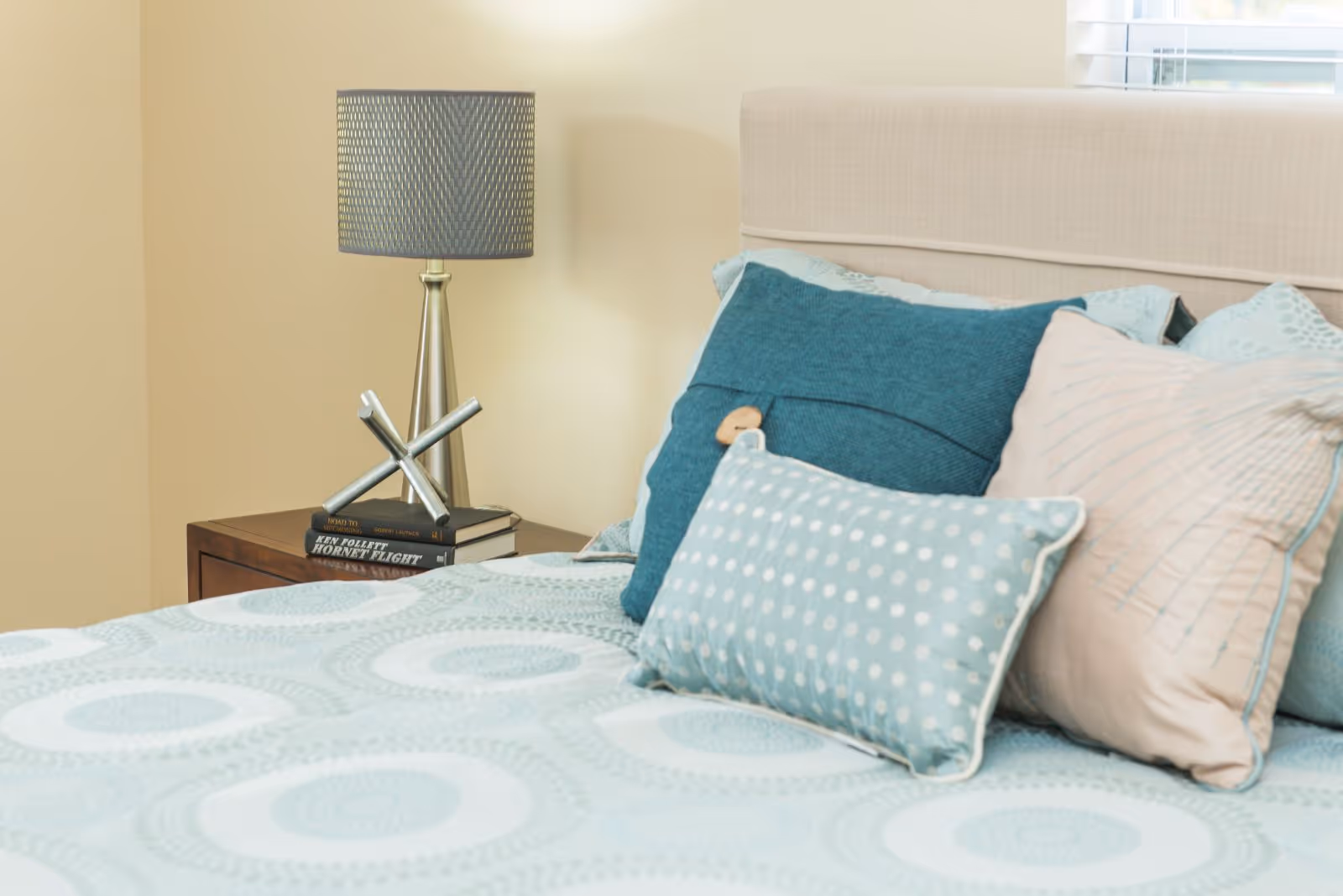 Close-up view of a bed with a beige upholstered headboard, decorated with multiple pillows in shades of blue and beige. Next to the bed is a wooden nightstand with a modern silver lamp and two stacked books.