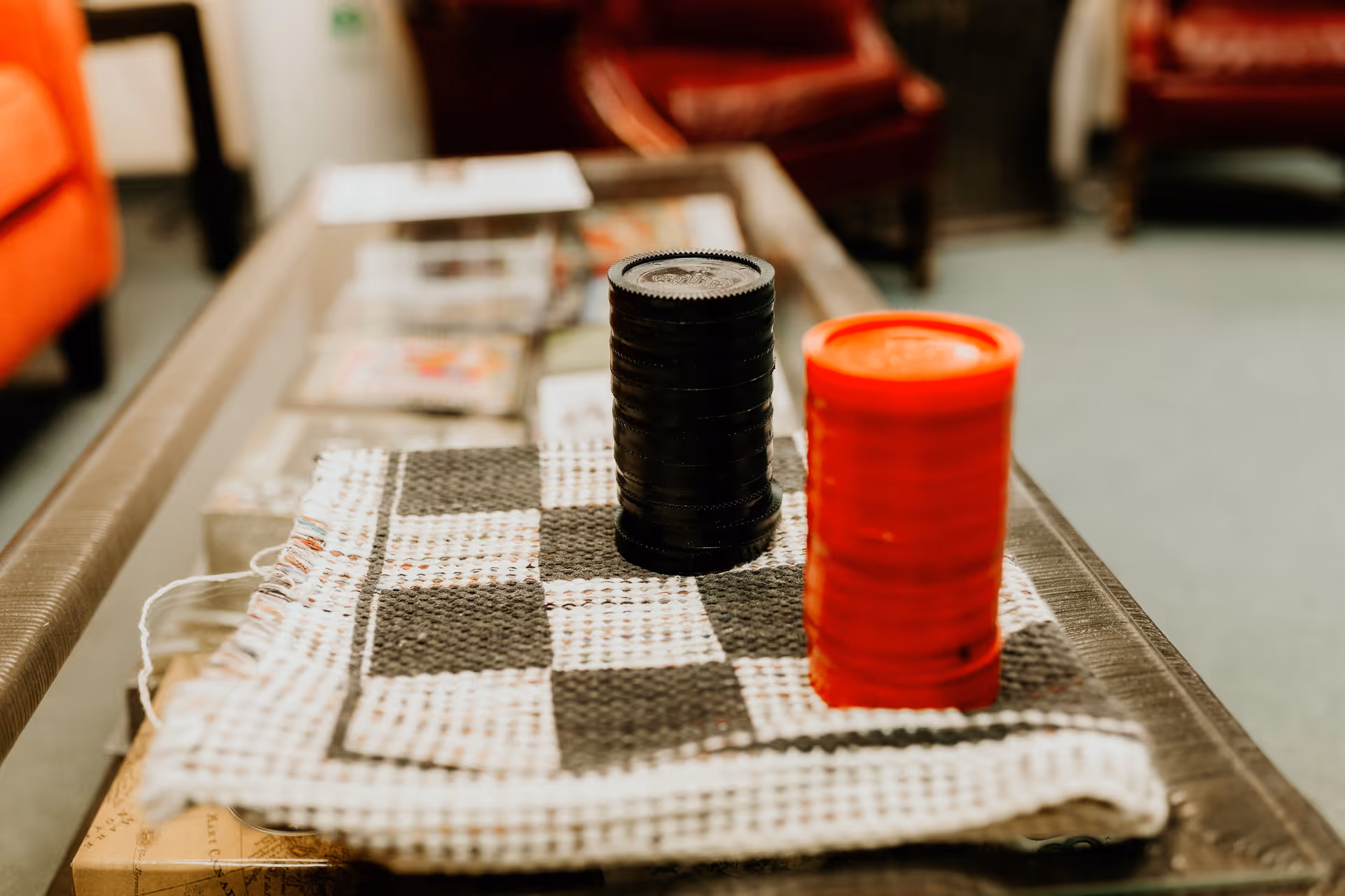 Two stacks of checkers pieces, one black and one red, placed on a black and white checkered cloth on a glass coffee table in a room with red armchairs and a carpeted floor.