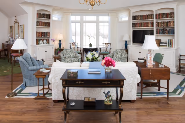 A bright and elegant living room with hardwood floors, white built-in bookshelves filled with books, and large windows letting in natural light. The room features a white sofa, two floral armchairs, a blue armchair, and several wooden tables with lamps and floral arrangements. A television is placed within one of the bookshelves.