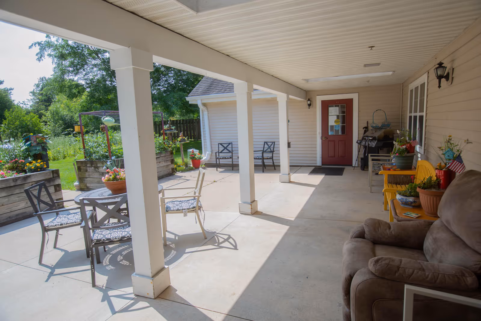 Covered outdoor patio area with several chairs and a table, potted plants, a red door, and a garden with raised flower beds in the background under a sunny sky.