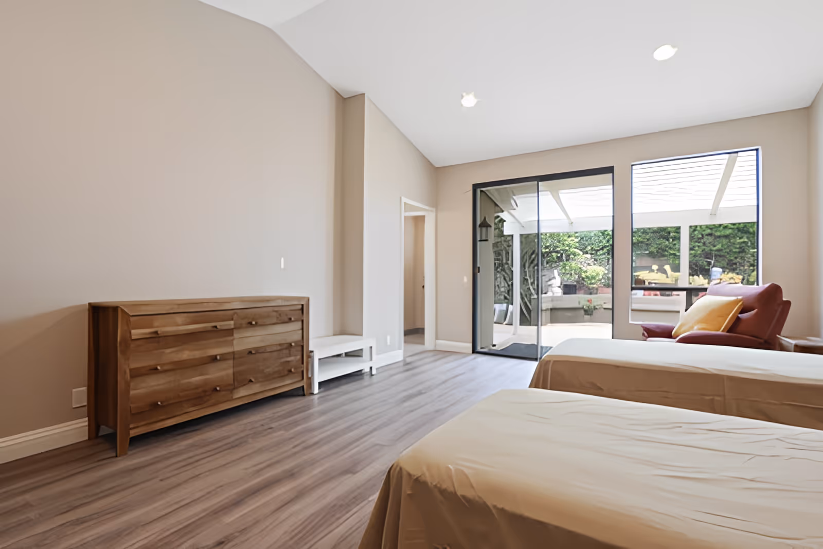 A bright and spacious bedroom with two beds covered in beige sheets, a wooden dresser against the left wall, a red armchair with a yellow pillow near a large window, and a sliding glass door leading to an outdoor patio area with greenery.