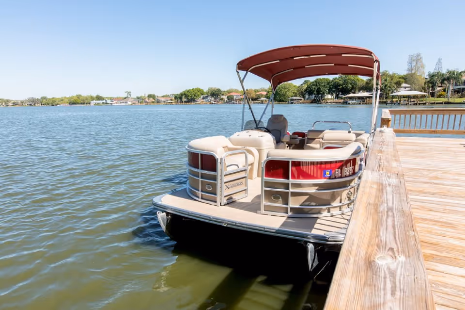 A pontoon boat with beige seats and a red canopy docked at a wooden pier on a calm lake with houses and trees visible in the background under a clear blue sky.