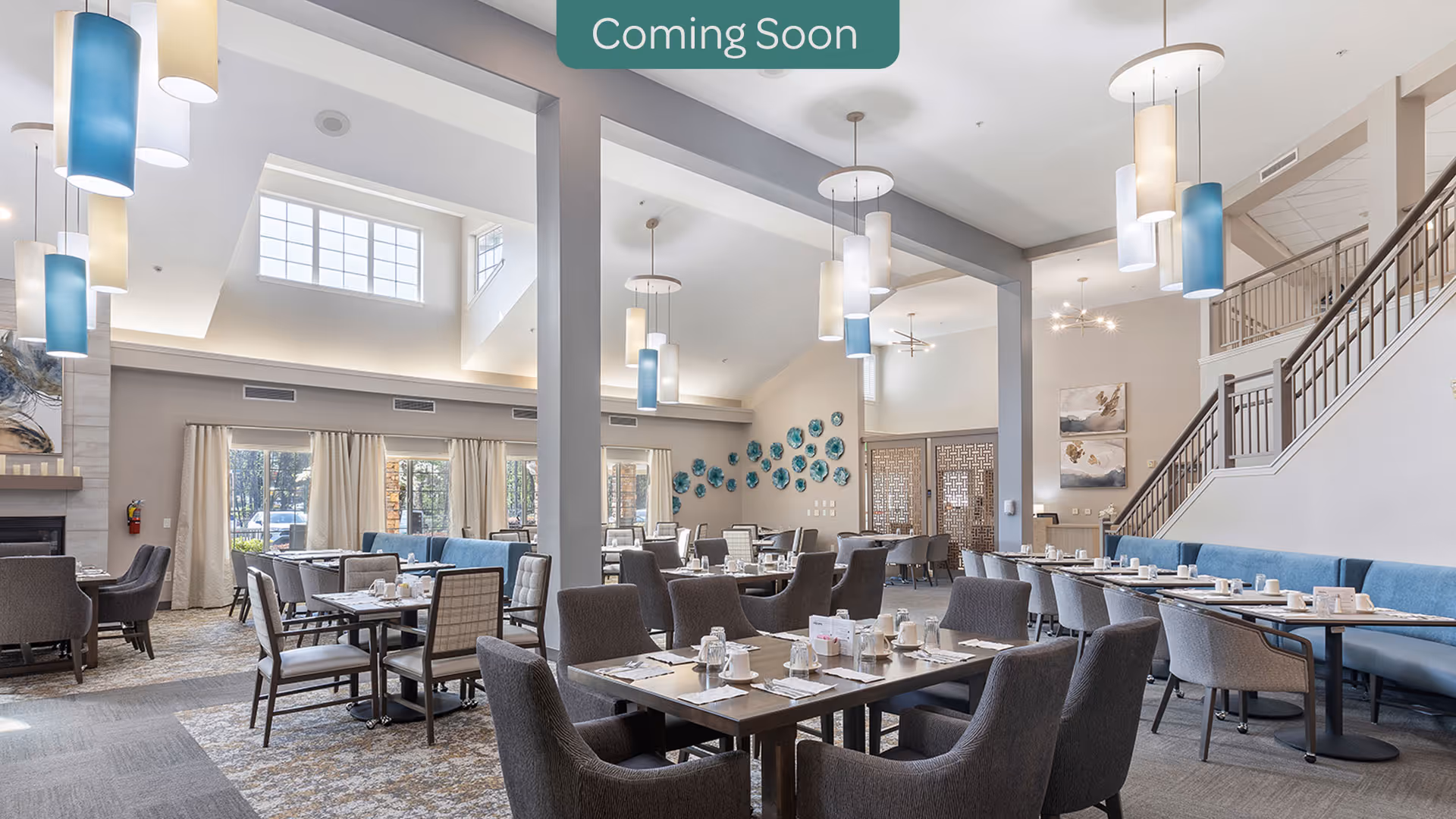 Bright, spacious dining room with multiple set tables and chairs, pendant lights, high ceilings, and a staircase.