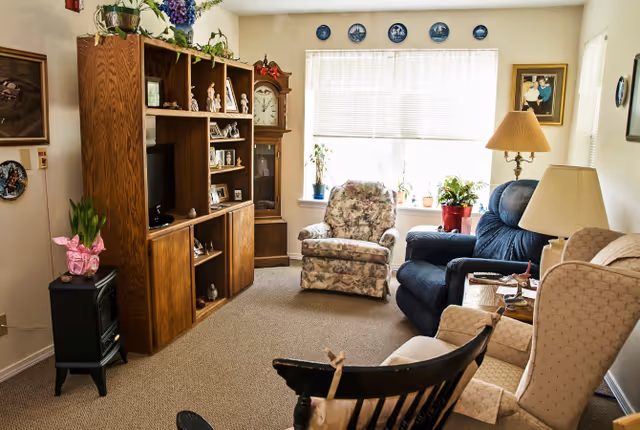 A cozy living room with a floral armchair, a blue recliner, and a beige wingback chair arranged around a wooden entertainment center with a TV and decorative items. There is a tall grandfather clock in the corner, a small black heater with a pink wrapped plant on top, and a window with blinds letting in natural light. The walls are decorated with framed pictures and decorative plates.