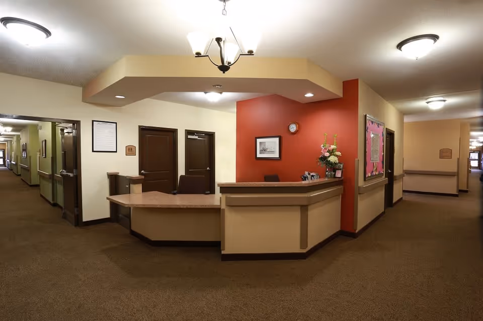 Reception desk area inside Gabriel Manor facility with a beige and red color scheme, a clock on the red wall, a framed picture, a flower arrangement on the desk, and hallways extending to the left and right.