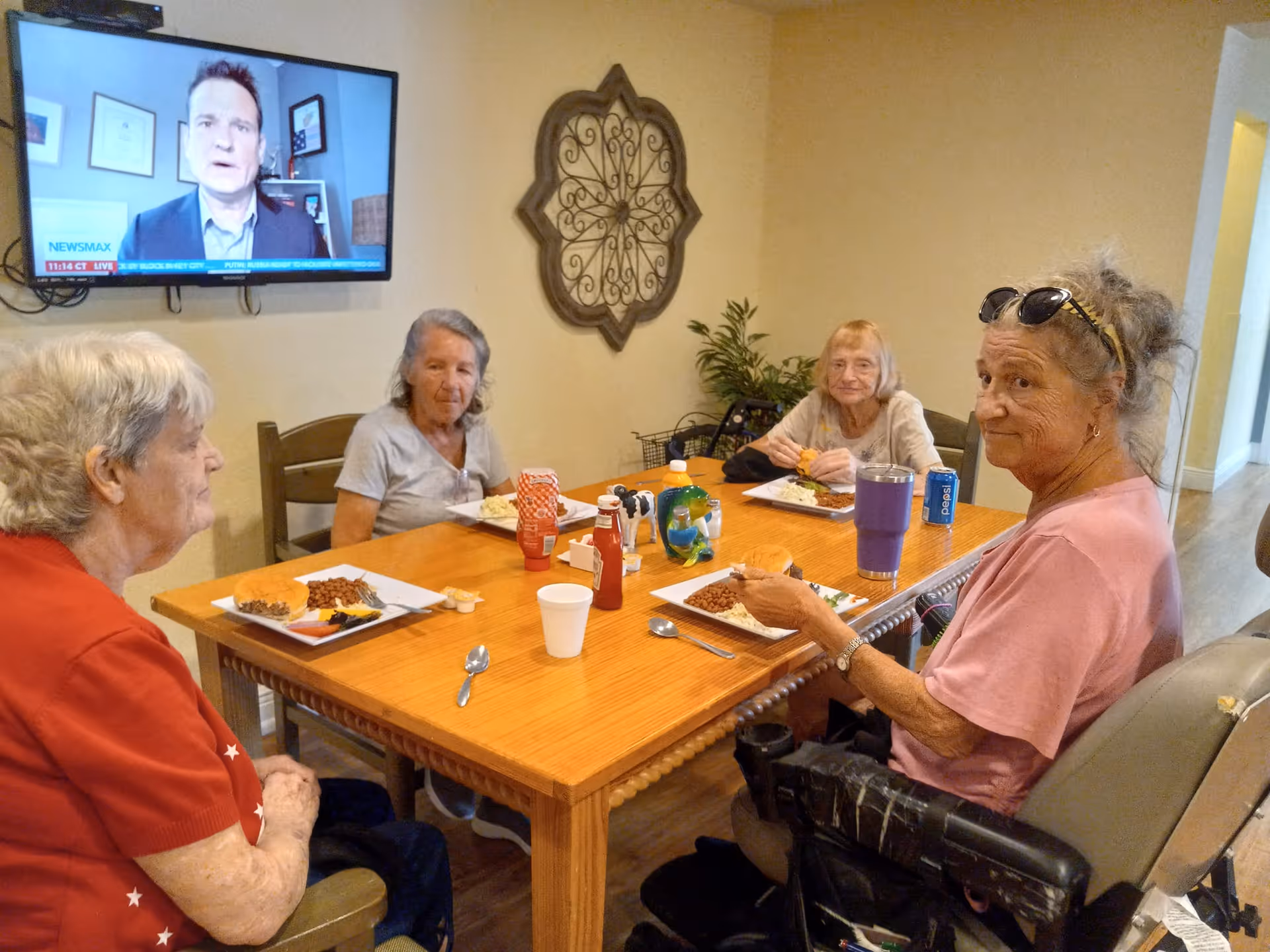 Four elderly women seated at a wooden dining table eating meals in a senior living facility common room with a TV on the wall.
