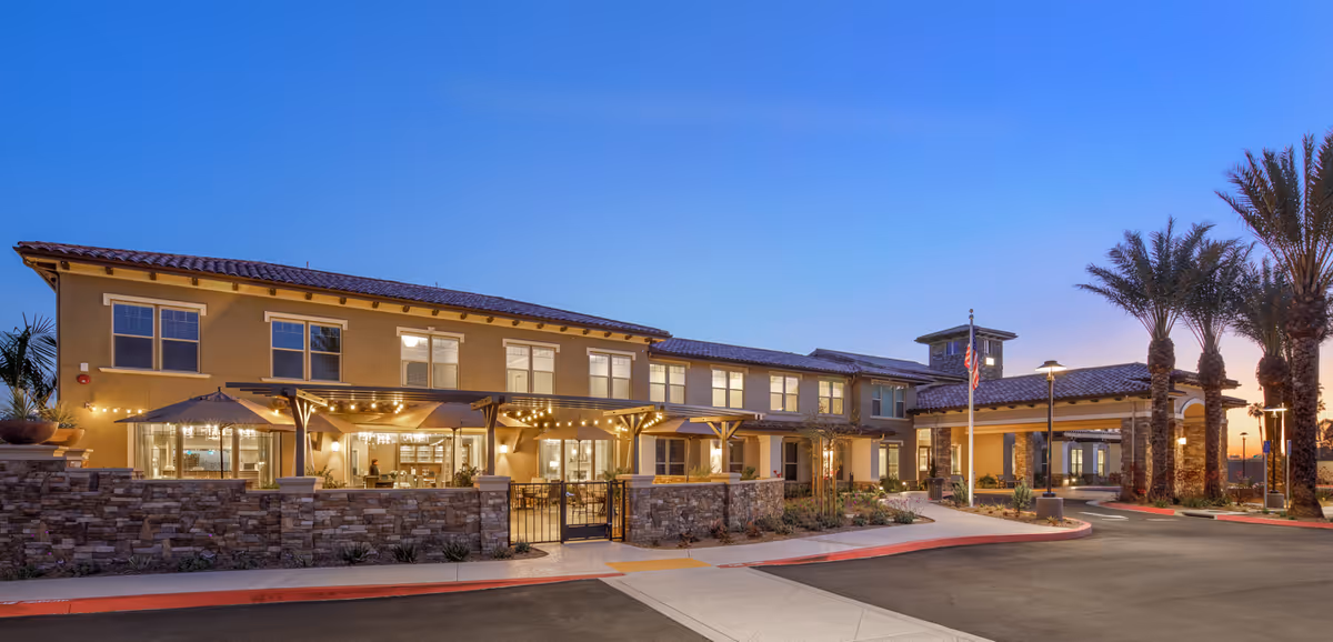 Exterior front entrance of a two-story senior living facility with stone walls, a lit patio, palm trees and a drive-up porte-cochère at dusk.