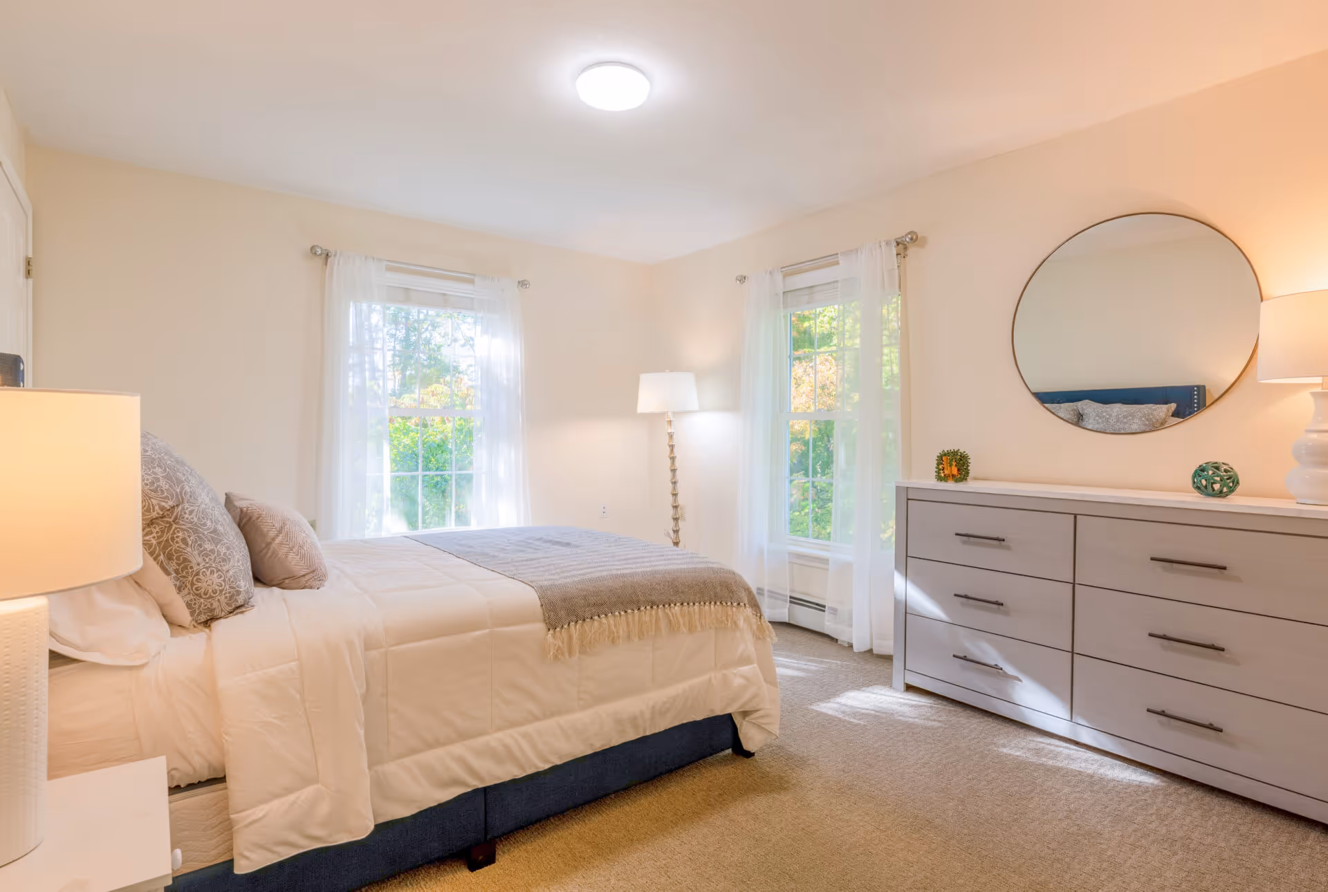 A bright and cozy bedroom with a neatly made bed featuring white and gray bedding, two windows with sheer white curtains allowing natural light to fill the room, a tall floor lamp in the corner, and a gray dresser with a round mirror and decorative items on top.