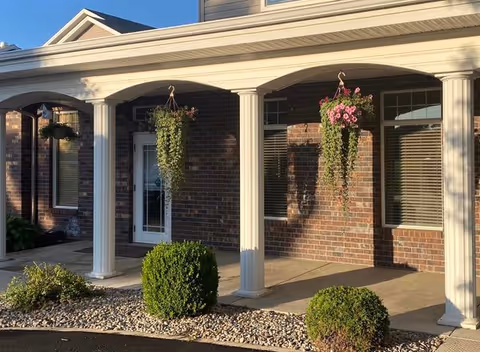 Front exterior view of a building with brick walls, white columns supporting a covered porch, hanging flower baskets, and neatly trimmed bushes along a stone-lined garden bed.