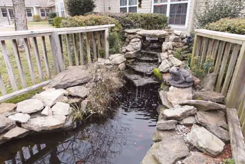 Rocky outdoor water feature with a small waterfall and wooden railing.