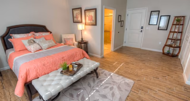 A bright and spacious bedroom with a large bed featuring coral and gray bedding, a tufted bench at the foot of the bed with a tray holding a plant and cups, a side table with a lamp, framed artwork on the wall, a cozy chair, and a wooden floor with a patterned rug. There is an open doorway leading to a bathroom and a white door on the far wall, along with a decorative wooden shelf holding plants and decor items.