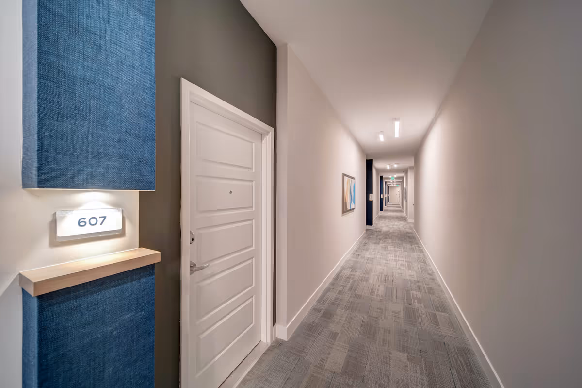 Long modern interior hallway with a white apartment door marked 607, carpeted floor, and recessed lighting.