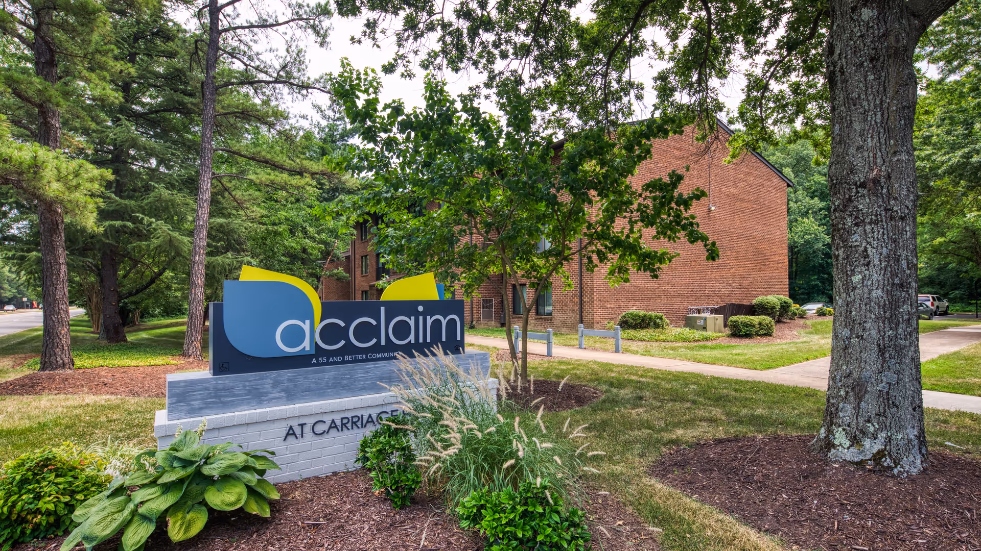 Outdoor view of Carriage Hill Apartments featuring a large sign with the word 'acclaim' and the phrase 'A 55 and better community' surrounded by trees, bushes, and grass. A brick apartment building is visible in the background along with a sidewalk and parked cars.