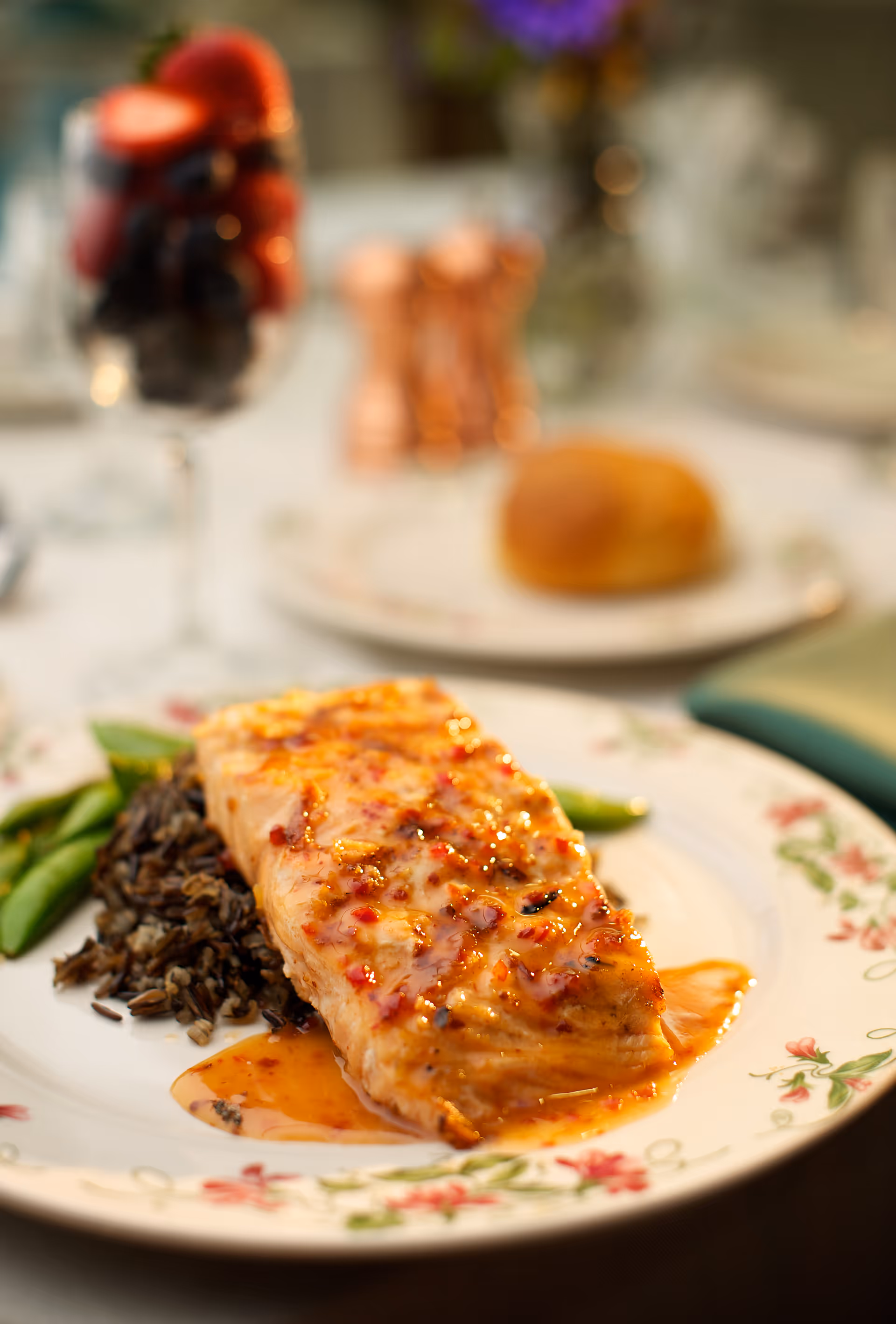 A plated meal featuring a piece of glazed salmon served on a bed of wild rice with green beans on the side. The plate has a floral pattern. In the background, there is a glass with berries and a bread roll on a separate plate, all set on a table with a white tablecloth.