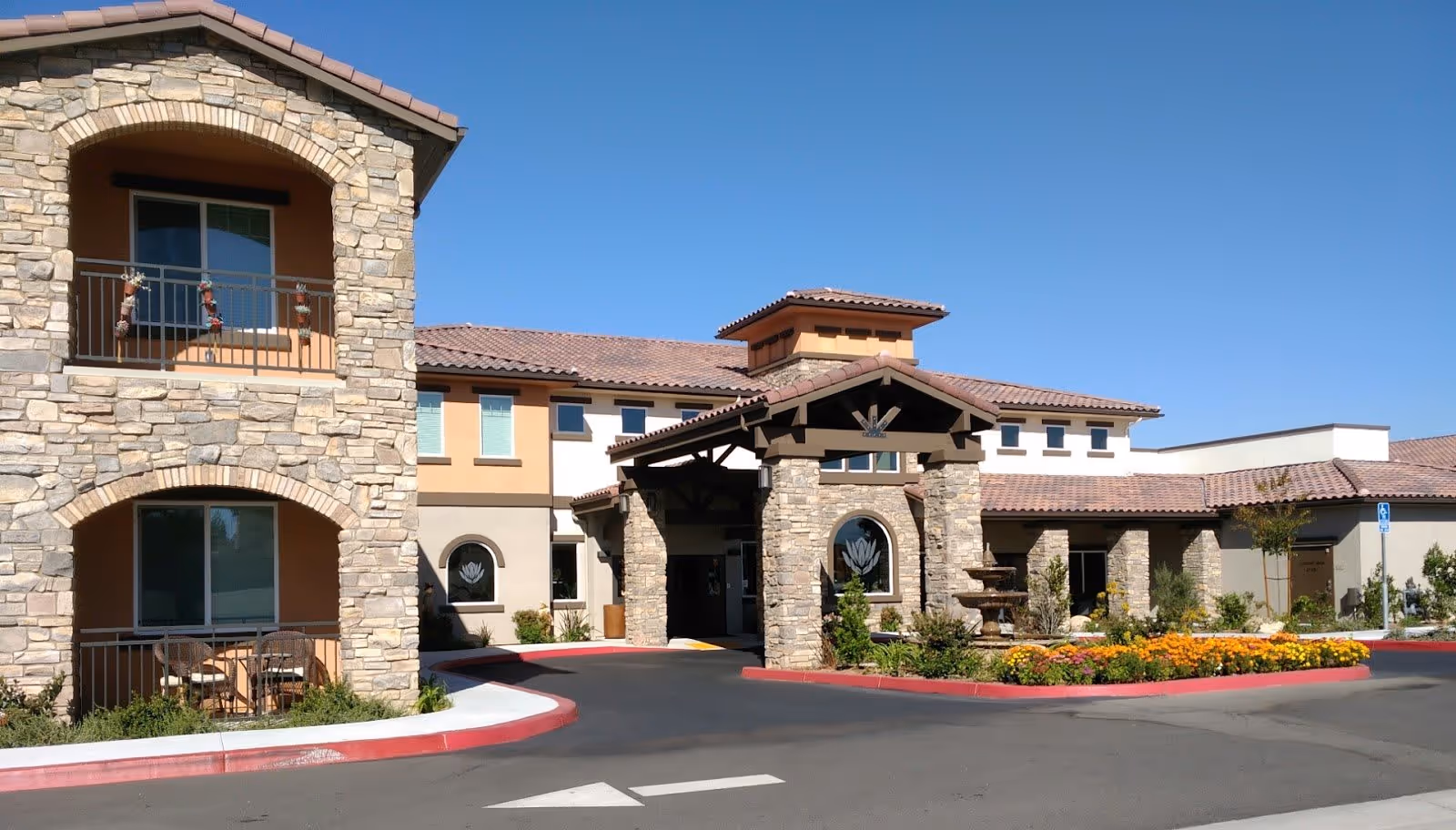 Exterior view of a senior living facility with stone and stucco architecture, featuring a covered entrance with stone pillars, a fountain, landscaped flower beds, and a clear blue sky.