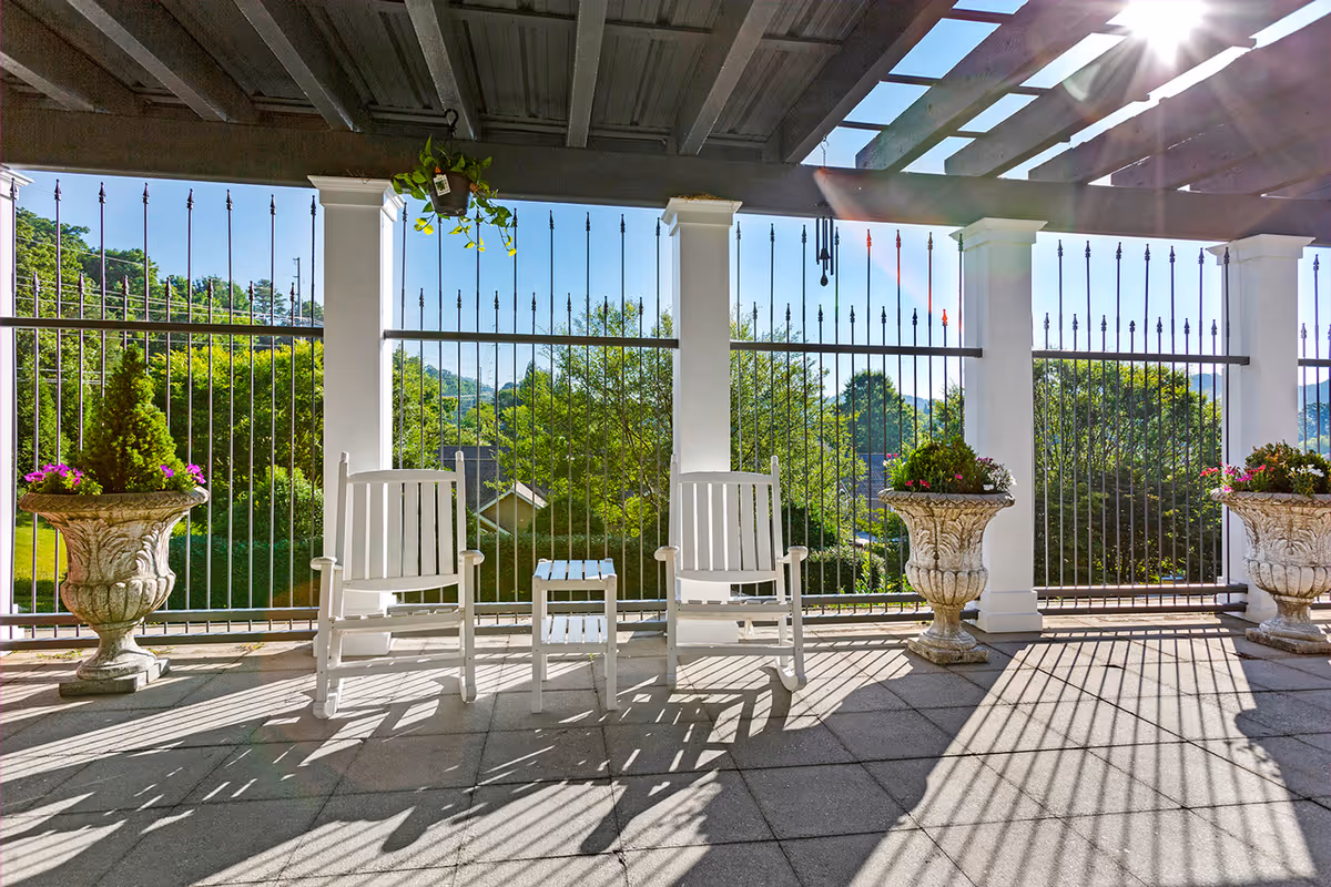 Sunlit covered patio with two white rocking chairs, a small side table, decorative urn planters, and a metal railing overlooking green trees.