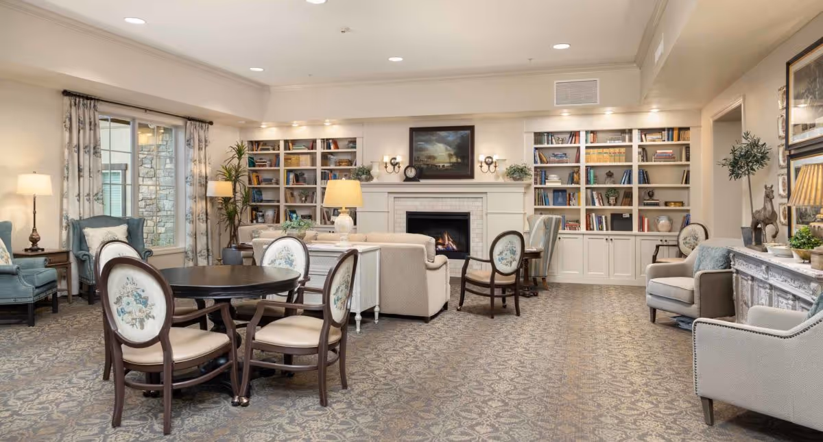 Well-lit communal living room with upholstered seating, a round table and chairs, a fireplace, and built-in bookshelves.