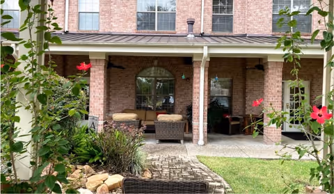 Outdoor covered patio area with wicker seating including a sofa and chairs, surrounded by green plants and flowers, attached to a brick building with large windows.