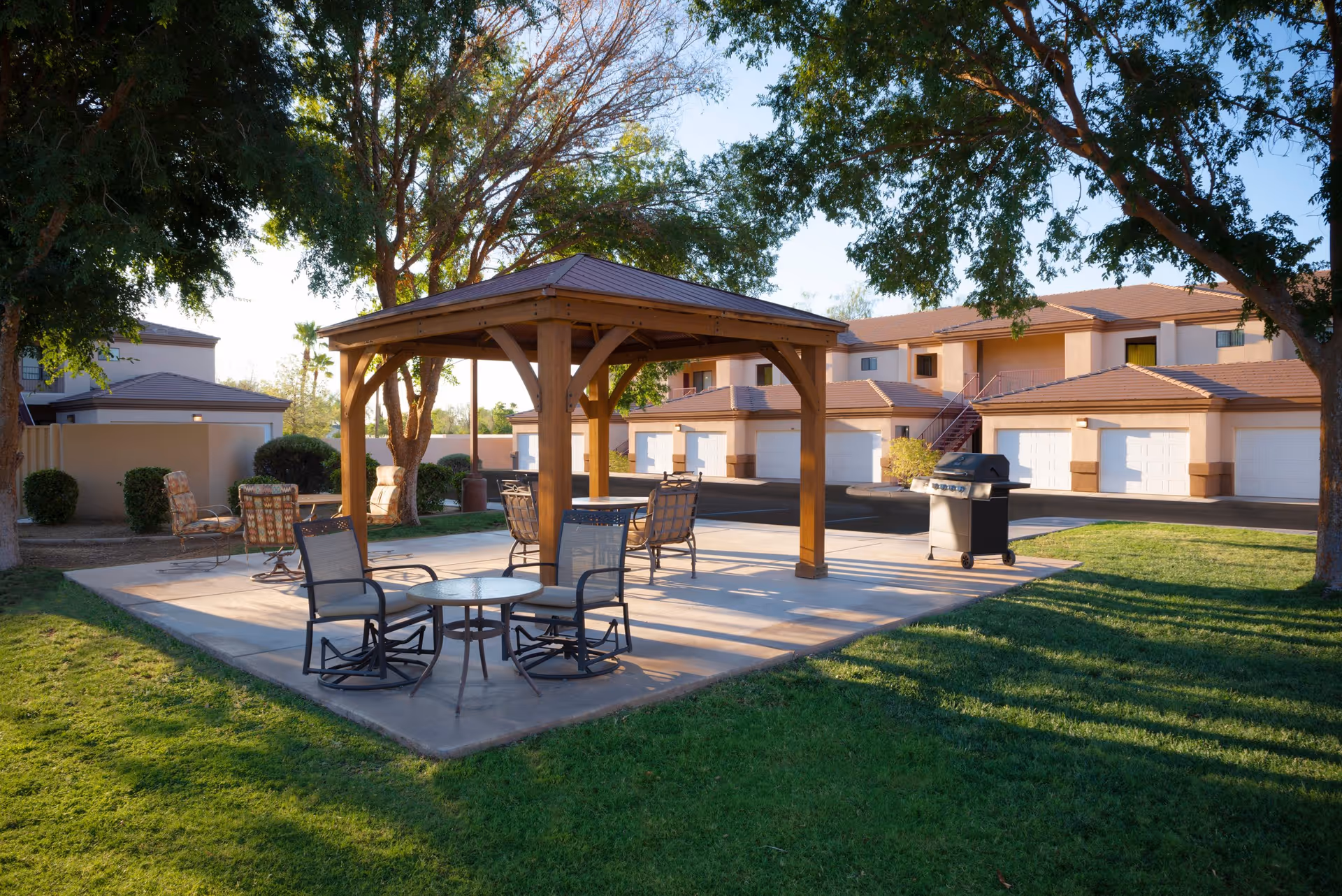 Outdoor seating area with a wooden gazebo, several chairs, tables, and a barbecue grill on a concrete patio surrounded by green grass and trees, with a background of residential buildings and garages under a clear sky.