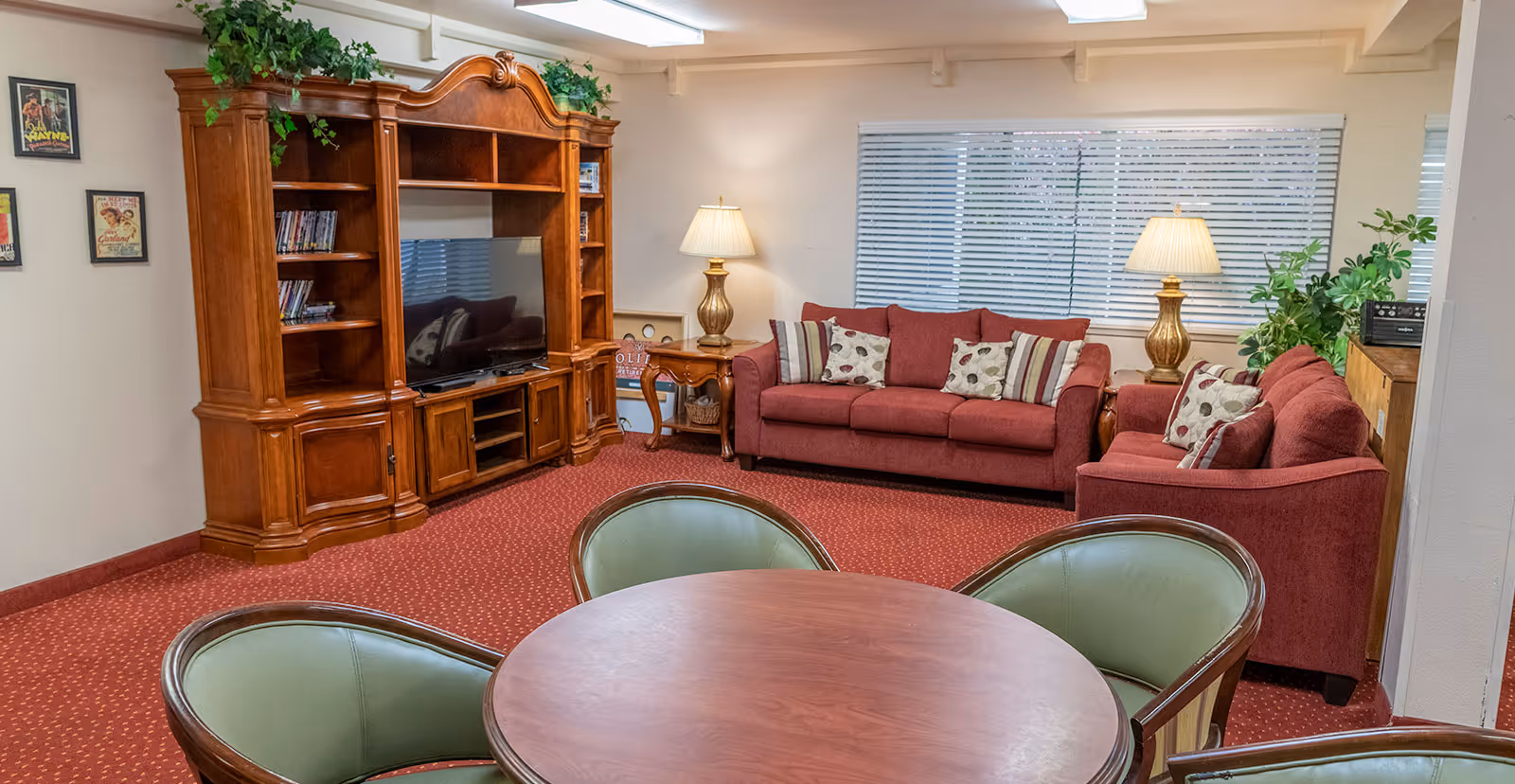 Common living area with red upholstered sofas, a wooden entertainment center with TV, a round table with green chairs, and lamps.