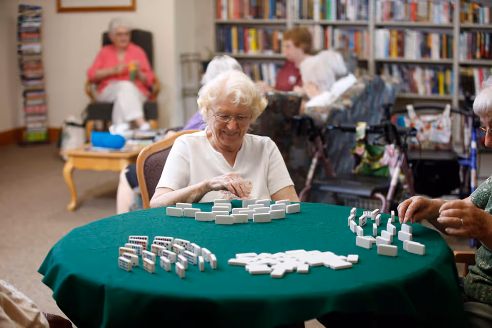 Elderly women sitting around a green table playing a game of dominoes in a cozy room with bookshelves and comfortable chairs in the background.