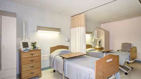 Two hospital-style beds with curtains and furniture in a shared bedroom.