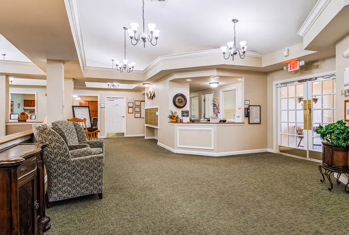 Interior view of a senior living facility lobby area with a reception desk, two patterned armchairs, a wooden side table, green carpet, beige walls, ceiling chandeliers, and glass double doors leading outside.