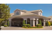 Front entrance of a beige senior living facility with a covered porte-cochère, columns, landscaped shrubs, and clear blue sky.