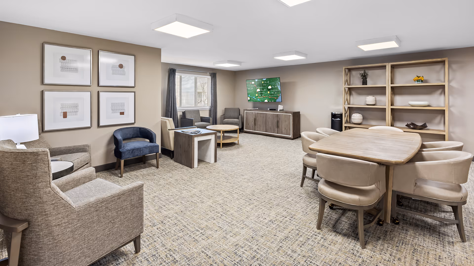 A spacious, well-lit common area in a senior living facility with beige walls and carpeted floor. The room features a wooden table surrounded by six beige cushioned chairs on the right, a wooden shelving unit with decorative items against the wall, and a flat-screen TV mounted above a wooden cabinet. On the left side, there are several armchairs in neutral tones and a small side table with a lamp. Four framed abstract artworks hang on the left wall, and a window with dark curtains lets in natural light.