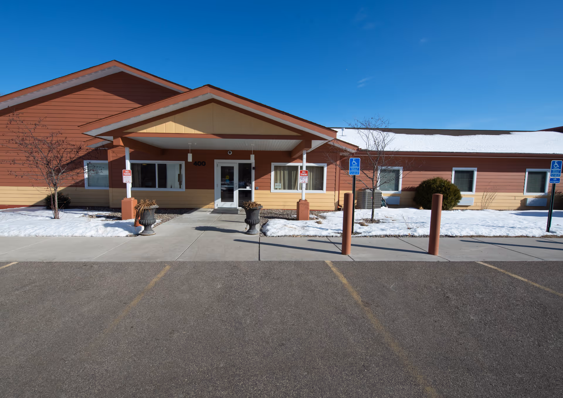 Front exterior view of a single-story building with a covered entrance, two large planters, and handicap parking spaces. Snow is visible on the ground and roof, under a clear blue sky.