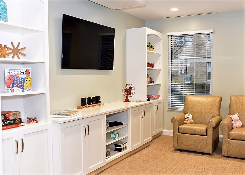 Cozy lounge with built-in white shelving and cabinets, a wall-mounted TV, and two upholstered chairs beside a window.