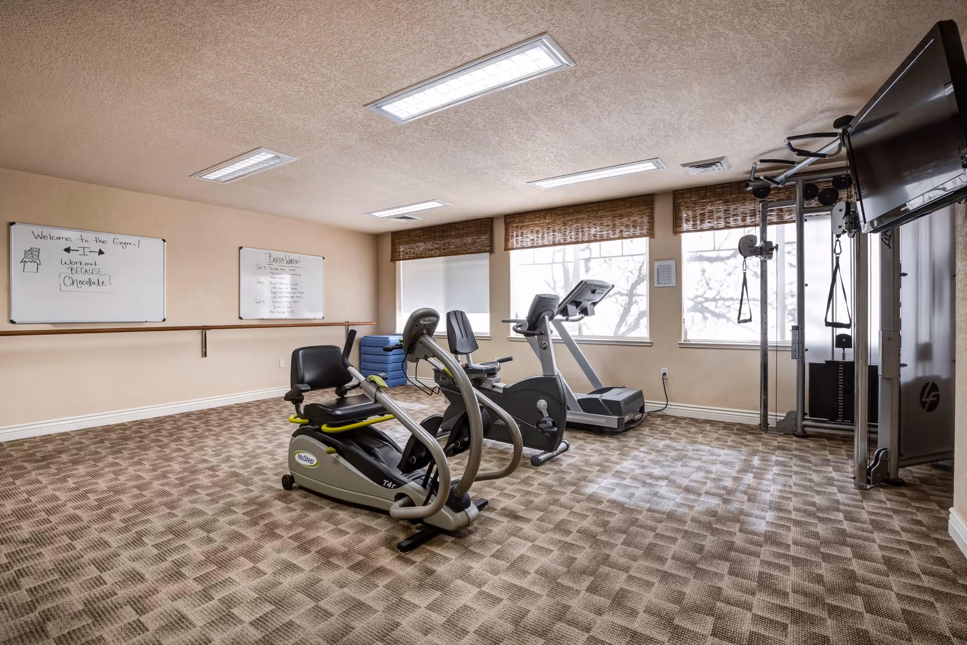 Small fitness room with exercise machines, a weight station, whiteboards on the wall, and windows with woven blinds.