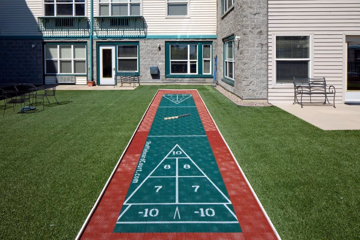 Outdoor shuffleboard court with scoring markings on a green and red surface, surrounded by artificial grass. The court is located in a courtyard area of a residential building with beige and gray exterior walls, windows, and patio furniture including benches and a table with chairs.