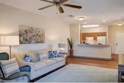 Bright open-plan living room with a sofa, armchair, ceiling fan, rug, wall art, and a view into a kitchen with a breakfast bar.