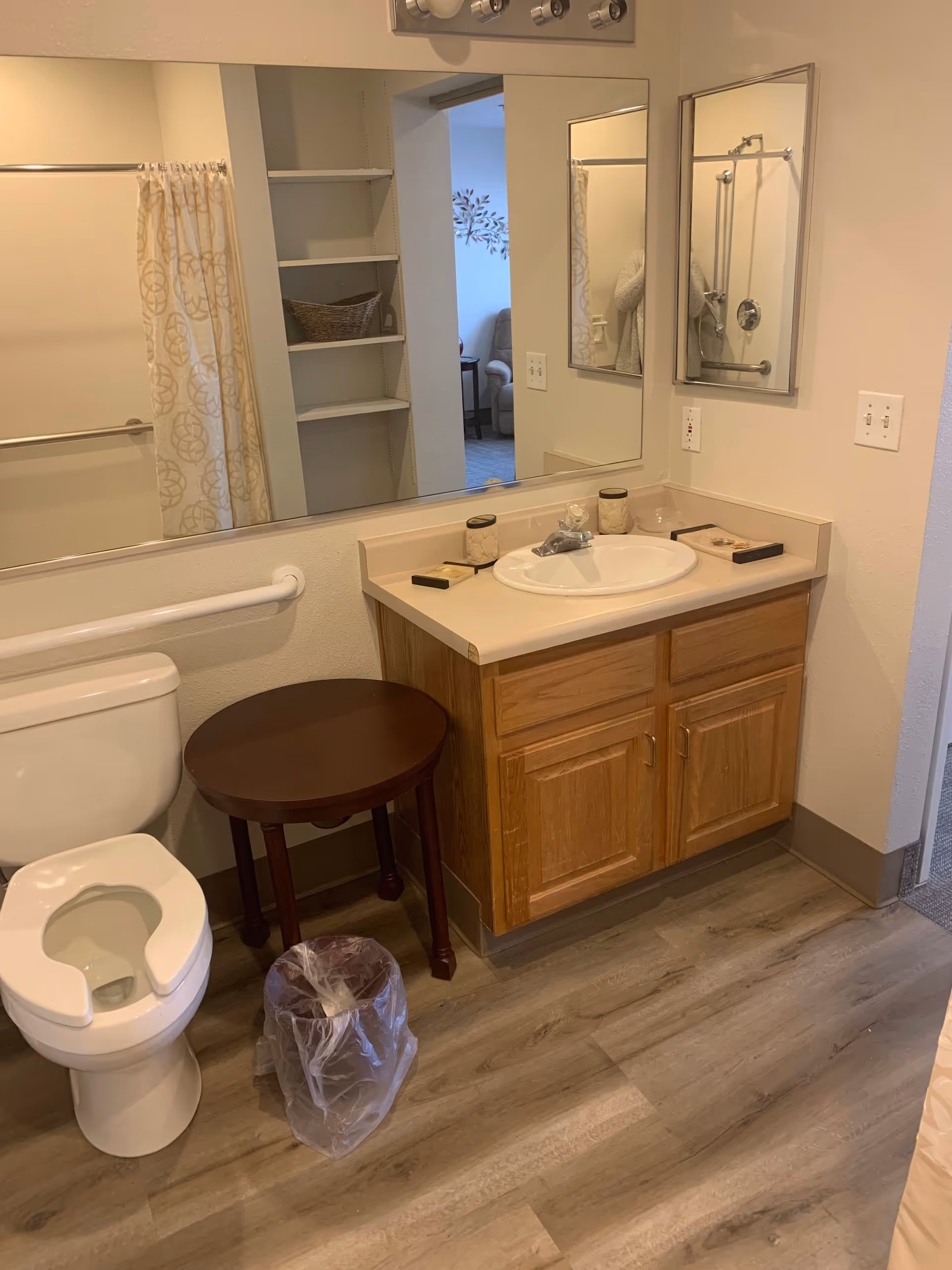 A bathroom with a toilet, a small round wooden table, a trash can with a plastic liner, a sink with a countertop and wooden cabinet below, a large mirror above the sink, and a shower curtain partially visible in the reflection. The floor has wood-like vinyl planks.