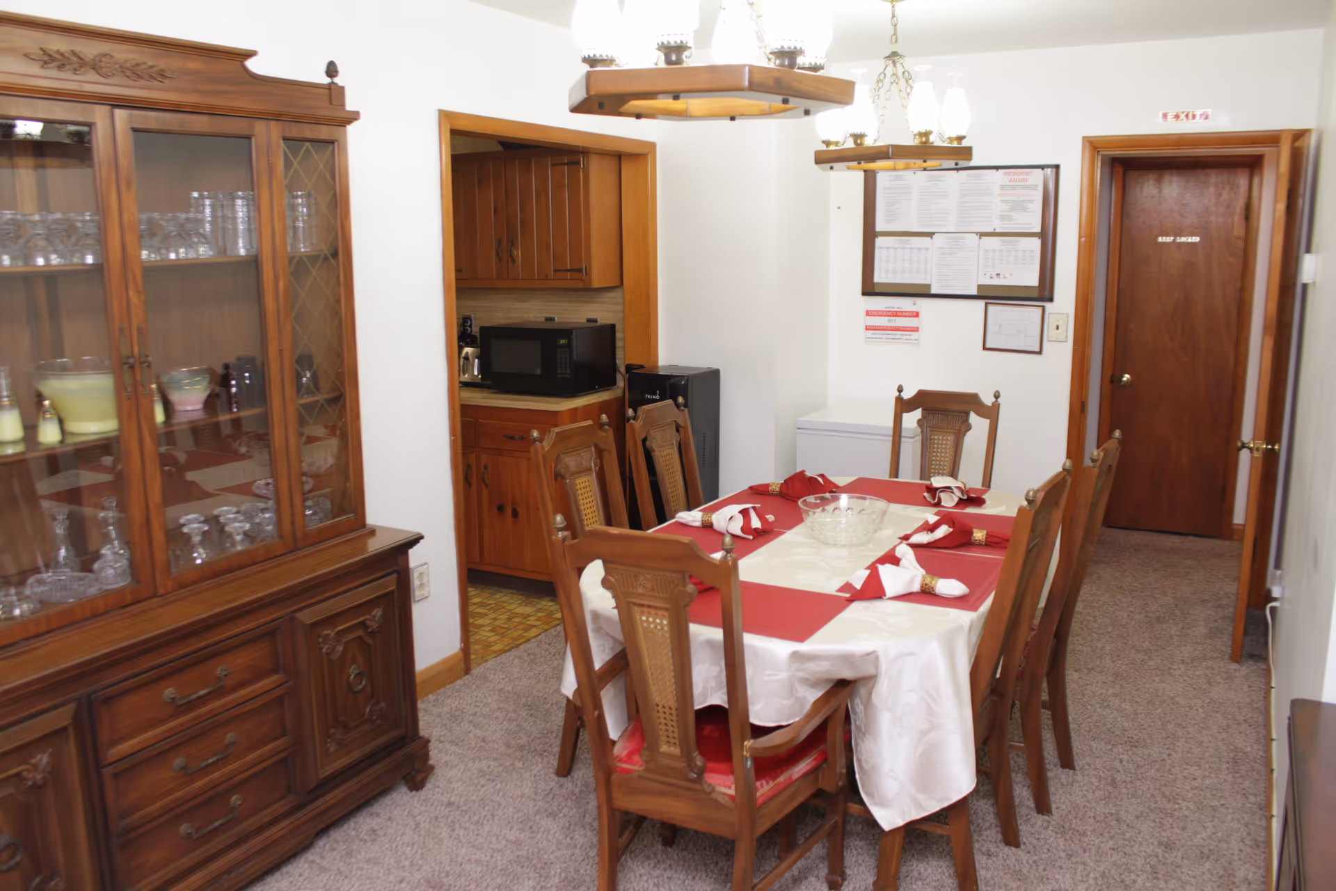 A dining room with a wooden table set with red placemats and napkins, wooden chairs, a china cabinet, and a doorway to a small kitchen.