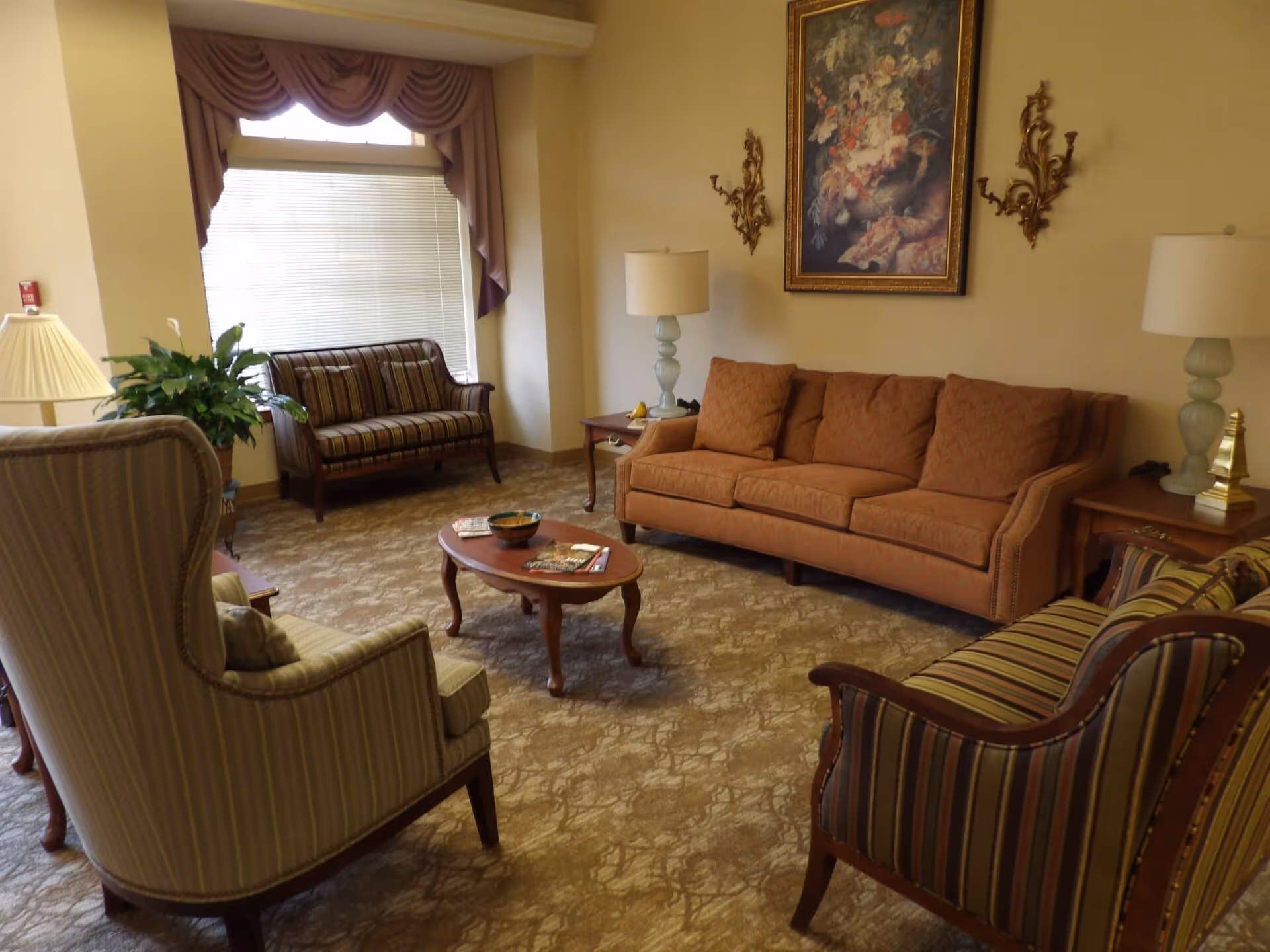 A cozy living room with a sofa, striped armchairs, a coffee table, lamps, framed artwork, and a window with drapery.
