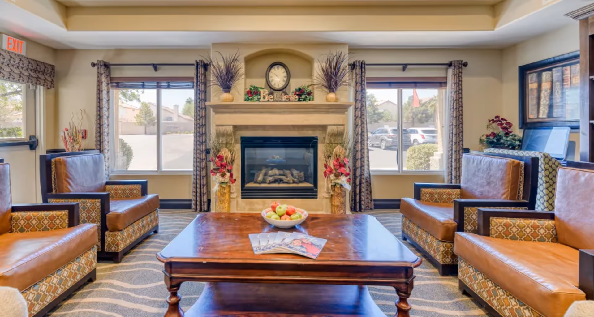 A cozy living room area with four leather and patterned armchairs arranged around a wooden coffee table. On the table is a bowl of apples and some magazines. The room features a fireplace with decorative plants and a clock above it. Two large windows with patterned curtains let in natural light, and there is an exit sign visible on the left side.