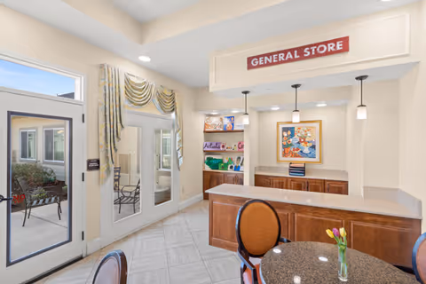 Interior view of a general store area in a memory care community with a counter, wooden cabinets, a round table with chairs, and a small vase with tulips. There is a sign above the counter that reads 'GENERAL STORE'. Glass doors and windows show an outdoor patio with chairs and plants.