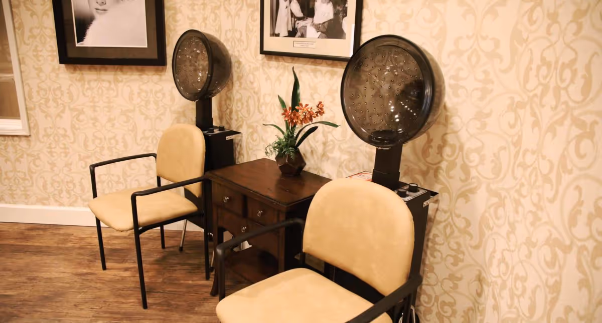 Two beige salon chairs with black armrests positioned in front of two vintage hair dryers mounted on stands. Between the chairs is a small wooden table with drawers, decorated with a potted plant with orange flowers. The wall behind has a beige patterned wallpaper and framed black and white photographs.