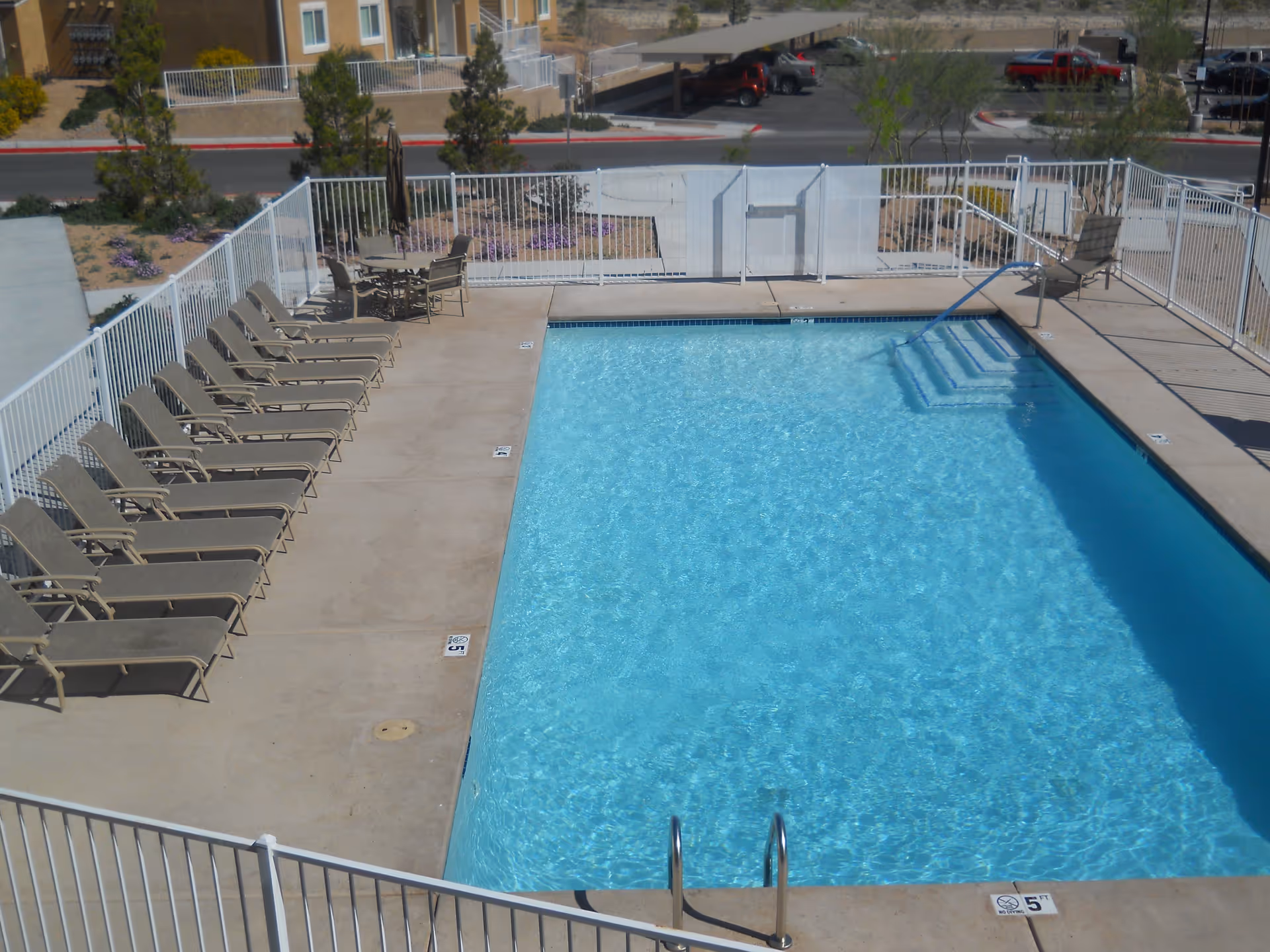 Outdoor swimming pool with clear blue water surrounded by a concrete deck. Several lounge chairs are lined up on one side, and a table with chairs and an umbrella is visible near the back. The pool area is enclosed by a white metal fence, and there are buildings and parked cars in the background.