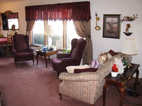 A cozy living room with a beige patterned sofa, two purple armchairs, a wooden side table with a lamp, and a window with burgundy and beige curtains. The room is decorated with framed artwork, a small statue, and various decorative items on the tables.