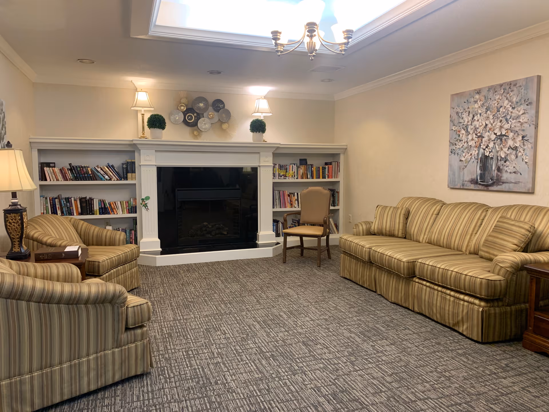 A cozy living room with striped upholstered armchairs and a matching sofa arranged around a white fireplace with built-in bookshelves on either side. The shelves are filled with books, and the fireplace mantel is decorated with two small potted plants and two lamps. A decorative wall art piece hangs above the sofa, and a ceiling light fixture illuminates the room.