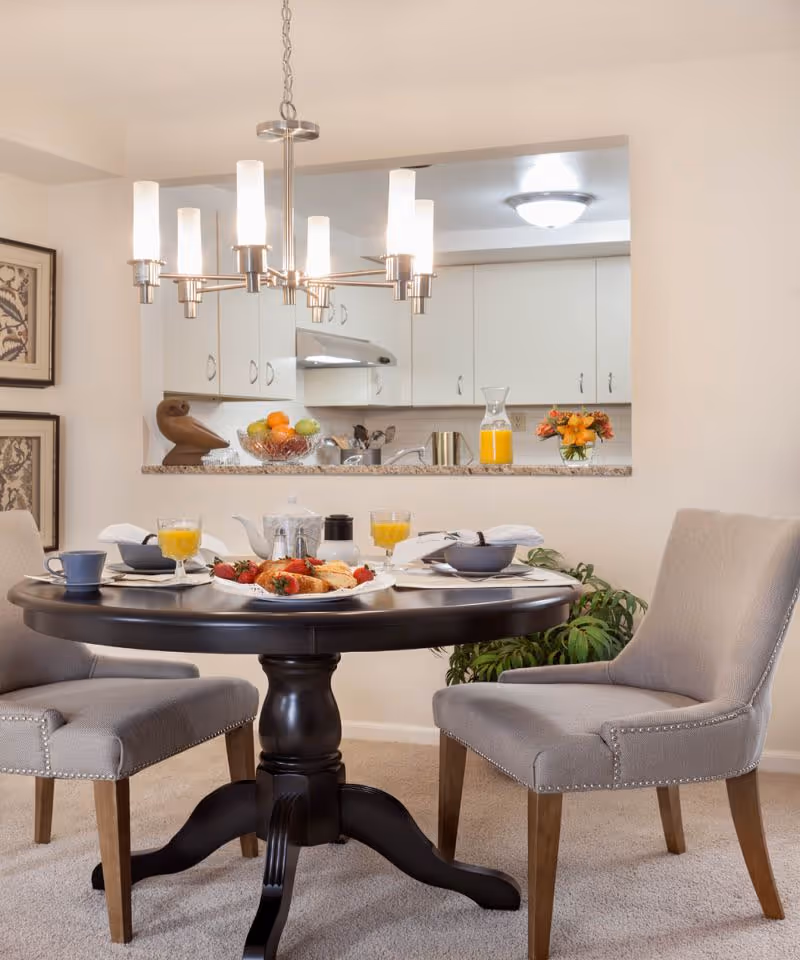 A cozy dining area with a round dark wooden table set for breakfast with plates of food, glasses of orange juice, and cups. Two upholstered gray chairs with wooden legs are positioned around the table. In the background, there is a kitchen with white cabinets, a fruit bowl, a pitcher of orange juice, and a small vase of flowers on the counter. A modern chandelier with multiple lights hangs above the table.