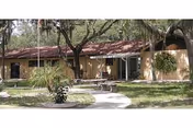 Single-story beige building with a curved path, trees, and residents near the entrance.