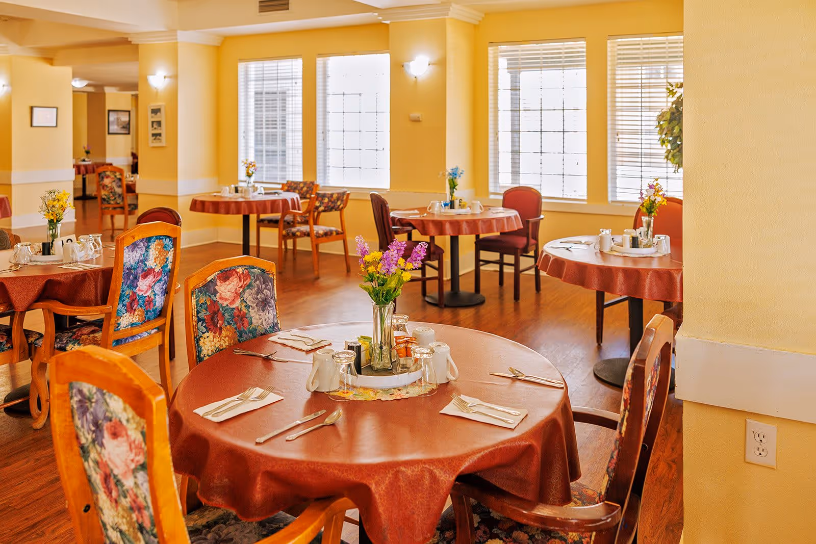 A bright dining room with round tables covered in red tablecloths, each set with utensils, napkins, cups, and small flower vases. The room has large windows with white blinds, yellow walls, and wooden flooring. Chairs with floral upholstery surround the tables.