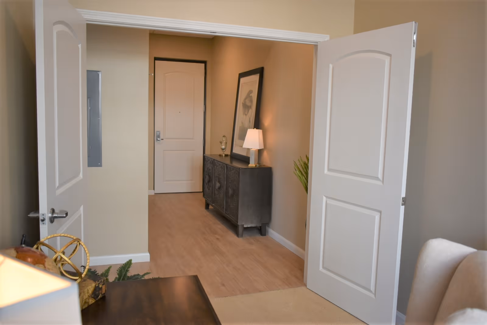 View through open double doors into a hallway with a closed white door at the end. The hallway has light wood flooring and beige walls. On the right side of the hallway, there is a dark console table with a lamp and a framed picture leaning against the wall. A small plant is visible next to the console table.