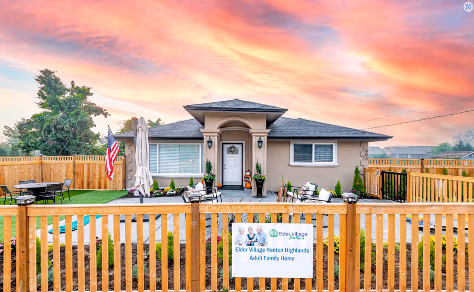 Front exterior view of a single-story adult family home with a fenced yard, outdoor seating, an American flag, and a colorful sunset sky in the background. A sign on the wooden fence reads 'Elder Village Renton Highlands Adult Family Home.'