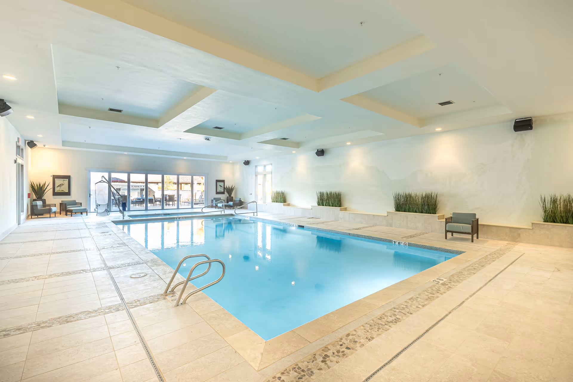 Indoor swimming pool area with light-colored tiled flooring and walls, recessed ceiling lights, several chairs and plants along the walls, and large glass doors leading to an outdoor patio with additional seating.